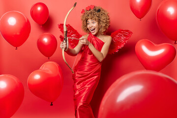 Happy positive female model pretends being cupid shoots with arrow at something wears red dress and...