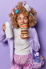 Vertical shot of funny pregnant woman holds pile of delicious sweet doughnuts and milk bottle has leaked makeup dressed in casual domestic clothes isolated over purple background. Happy expectations