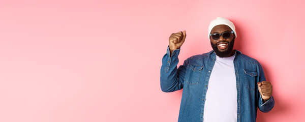 Happy Black man in beanie an sunglasses rejoicing, dancing with happy face, standing over pink background