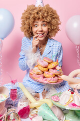 Happy surprised woman smiles broadly keeps finger in mouth holds delicious glazed doughnuts surrounded by tasty desserts poses near messy room isolated on pink background. Birthday celebration concept