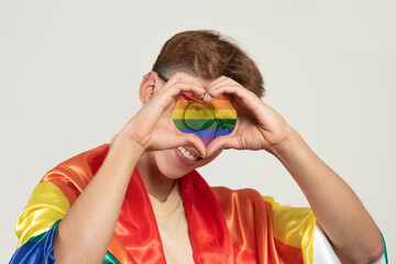 Happy young caucasian man covered with the flag of the lgtbqi community making the shape of a heart...