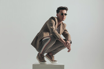 cool fashion model with plaid pants and beige jacket crouching on white box