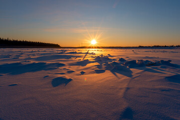 The bright sun at sunset sets behind a forest on the Vilyui River in Yakutia in winter against the backdrop of a forest with power lines and houses in the village of Suntar.
