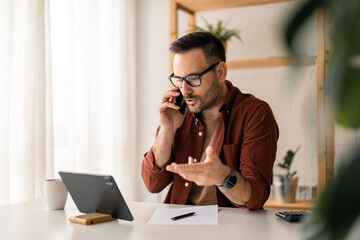 Dedicated young businessman wearing casual clothes and glasses holding smartphone, having a call in home office, working, consulting client, providing business service via mobile phone.