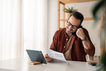 Smiling businessman, salesman talking on smartphone consulting client making call in home office working looking at digital tablet making loan offer, wearing business casual clothes, glasses and watch
