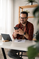 Fototapeta na wymiar Happy smiling businessman wearing casual clothes and using modern smartphone in his home office, successful employer sitting at desk typing on mobile phone, also using digital tablet, pen and paper.