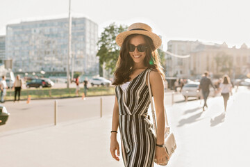 Pretty charming woman with dark hair and wonderful smile wearing summer suit, hat and sunglasses posing at camera during summer walking in the city on sunset.