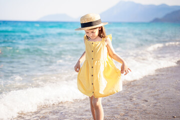 Little girl walking on beach, sea ocean shore in romantic yellow dress, straw hat. Playing in sand, blue waves. Family vacation travel leisure in hot summer coast. Sunny day relax in hotel resort