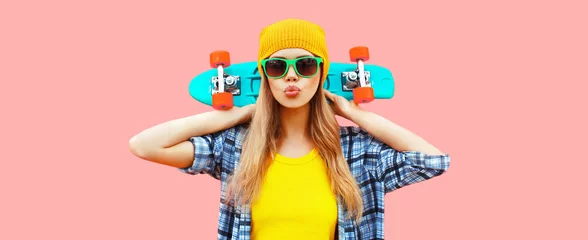 Tuinposter Portrait of stylish blonde young woman model with skateboard wearing colorful yellow hat on pink background © rohappy