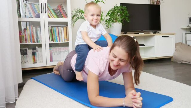 Cute baby boy sitting on mothers back while she is standing in plank during fitness training lesson. Concept of healthcare, sports and yoga at home