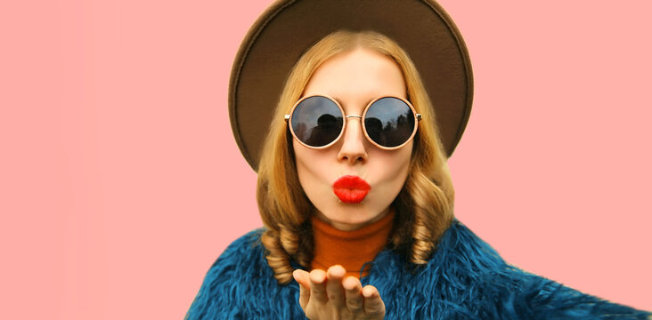 Fashionable portrait of stylish young woman stretching hand for taking selfie blowing her lips with red lipstick sending sweet air kiss on pink background