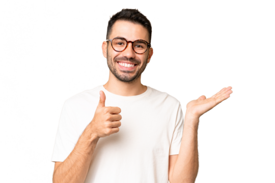 Young handsome caucasian man over isolated chroma key background holding copyspace imaginary on the palm to insert an ad and with thumbs up