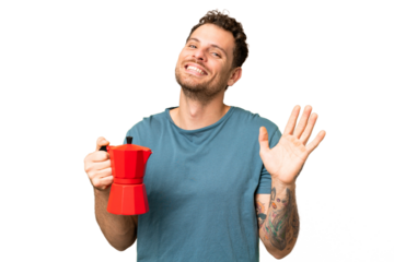 Brazilian man holding coffee pot over isolated chroma key background saluting with hand with happy expression