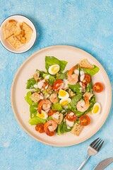 Caesar salad with romaine lettuce, cherry tomatoes, boiled quail eggs and croutons on a light ceramic plate on a blue concrete background. American cuisine.
