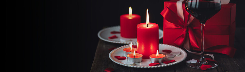 Obraz na płótnie Canvas Saint Valentine's Day celebration. Red burning candles, hearts, gift box, postcard on dark wooden background. Happy holiday. Table decor for festive dinner, romantic atmosphere. Banner copy space text