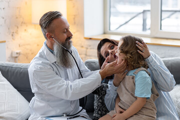 Kind male senior paediatrician doctor visiting his patient at home, examining little girl sitting on mother's lap, writing prescription. Concept of kid's health check