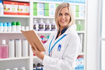 Young blonde woman pharmacist smiling confident reading notebook at pharmacy