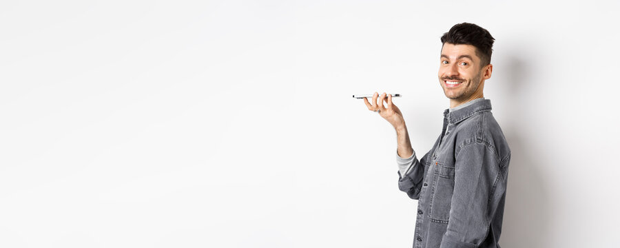 Profile Portrait Of Young Man Smiling At Camera, Recording Voice To Translate Word Or Leave Message, Talking On Speakerphone, Standing Against White Background