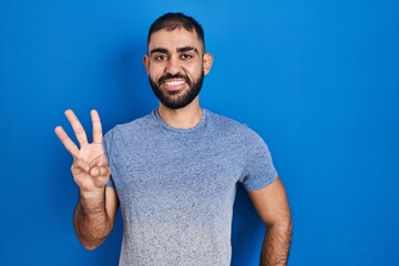 Middle east man with beard standing over blue background showing and pointing up with fingers number three while smiling confident and happy.