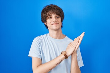 Hispanic young man standing over blue background clapping and applauding happy and joyful, smiling...