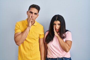 Naklejka premium Young couple standing over isolated background hand on mouth telling secret rumor, whispering malicious talk conversation