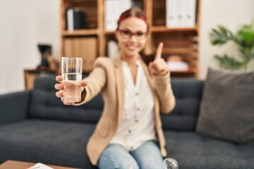Young caucasian woman offering a glass of water smiling with an idea or question pointing finger with happy face, number one