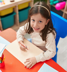 Adorable hispanic girl preschool student sitting on table drawing on paper at kindergarten