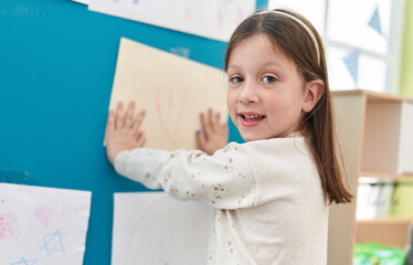 Adorable hispanic girl preschool student hanging heart draw on wall at kindergarten