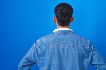 Fototapeta premium Young hispanic man standing over blue background standing backwards looking away with arms on body