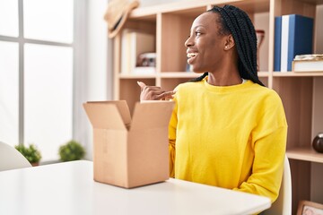 Beautiful black woman opening cardboard box pointing thumb up to the side smiling happy with open mouth