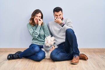 Young hispanic couple sitting on the floor with dog smelling something stinky and disgusting, intolerable smell, holding breath with fingers on nose. bad smell - obrazy, fototapety, plakaty