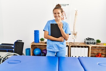 Young beautiful hispanic woman physiotherapist smiling confident holding document at rehab clinic