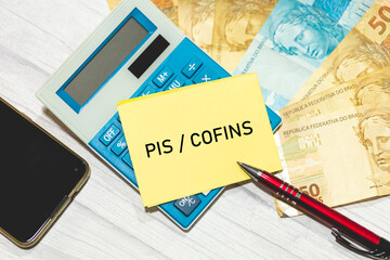 As siglas PIS e COFINS escritas em um pedaço de papel que está sobre uma calculadora. Notas do...
