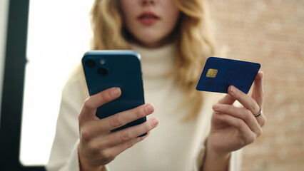 Young blonde woman using smartphone and credit card sitting on sofa at home