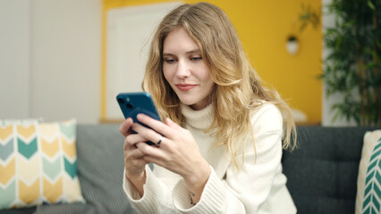 Young blonde woman using smartphone sitting on sofa at home