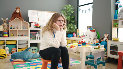 Young blonde woman preschool teacher stressed sitting on chair at kindergarten