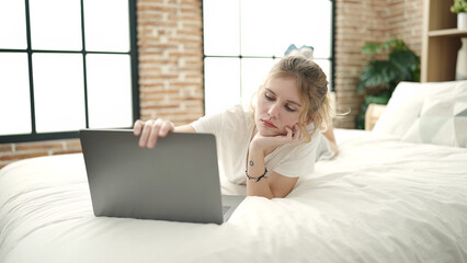 Young blonde woman using laptop lying on bed at bedroom