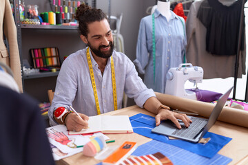 Young hispanic man tailor using laptop writing on notebook at atelier