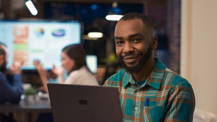 Smiling african american office employee working on laptop portrait. Young man holding computer, looking at camera close up, company financial presentation, business meeting background. Handheld shot.