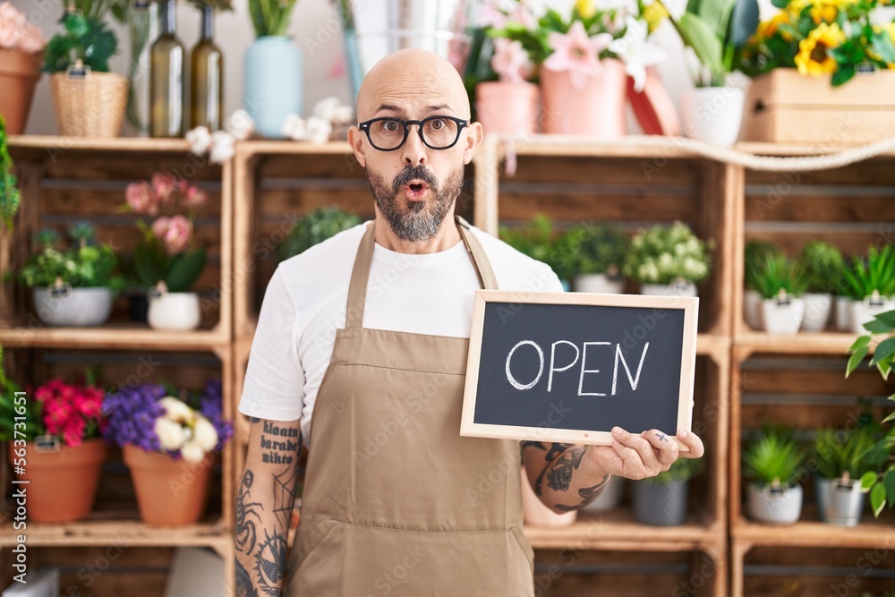 Sticker hispanic man with tattoos working at florist holding open sign scared and amazed with open mouth for