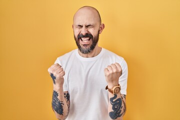 Young hispanic man with tattoos standing over yellow background very happy and excited doing winner...