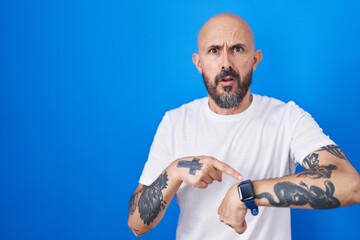 Hispanic man with tattoos standing over blue background in hurry pointing to watch time,...