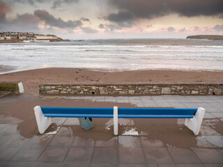 Blue color wooden bench by Kilkee beach and town houses in the background. County Clare, Ireland. Nobody. Popular summer resort. Atlantic ocean, Irish seascape. Cloudy sky.
