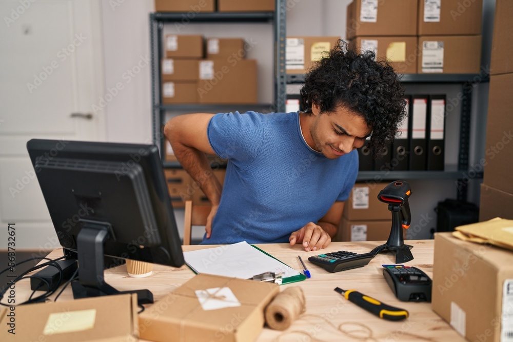 Poster Hispanic man with curly hair working at small business ecommerce suffering of backache, touching back with hand, muscular pain