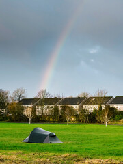 Simple tent on green grass in a park. Row of houses in the background. Rainbow in a blue sky. Homeless living close to dense populated area. Social issue.