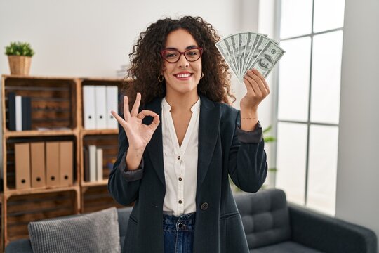 Young hispanic woman holding dollars doing ok sign with fingers, smiling friendly gesturing excellent symbol