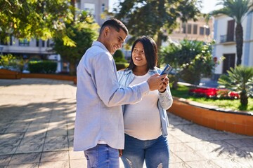 Fototapeta premium Young latin couple expecting baby using smartphone at park