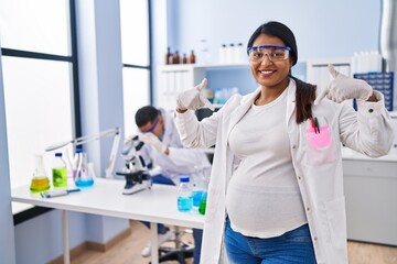 Young hispanic woman expecting a baby working at scientist laboratory looking confident with smile on face, pointing oneself with fingers proud and happy.