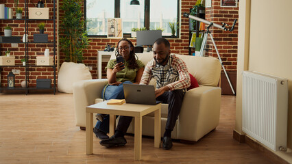 African american people relaxing at home on sofa, scrolling through social media app on smartphone. Enjoying free time leisure activity and laughing together, cozy recreation in living room.
