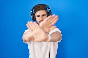 Hispanic man with beard listening to music wearing headphones rejection expression crossing arms and palms doing negative sign, angry face